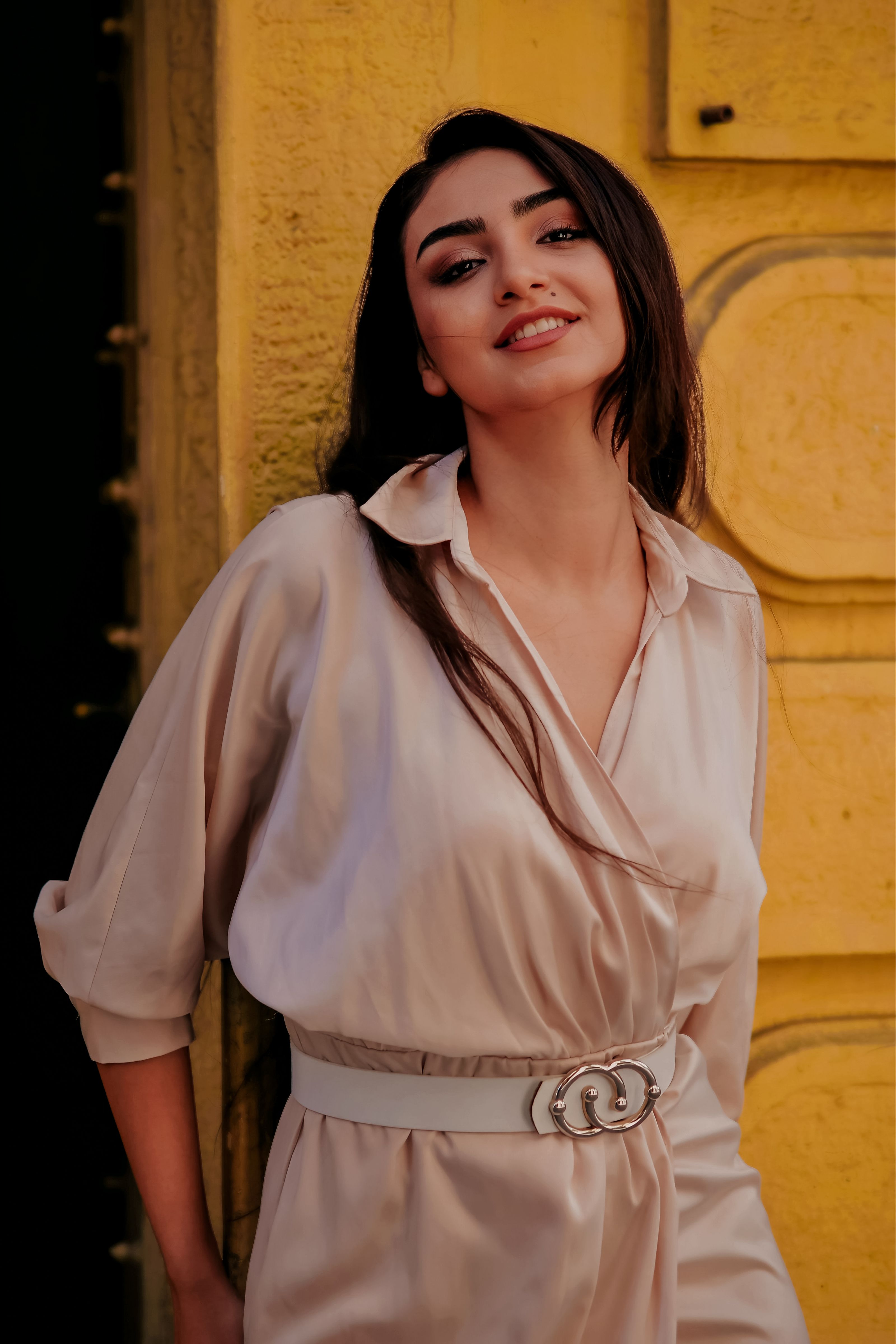 Mulher elegante de vestido bege sorrindo contra um fundo amarelo.