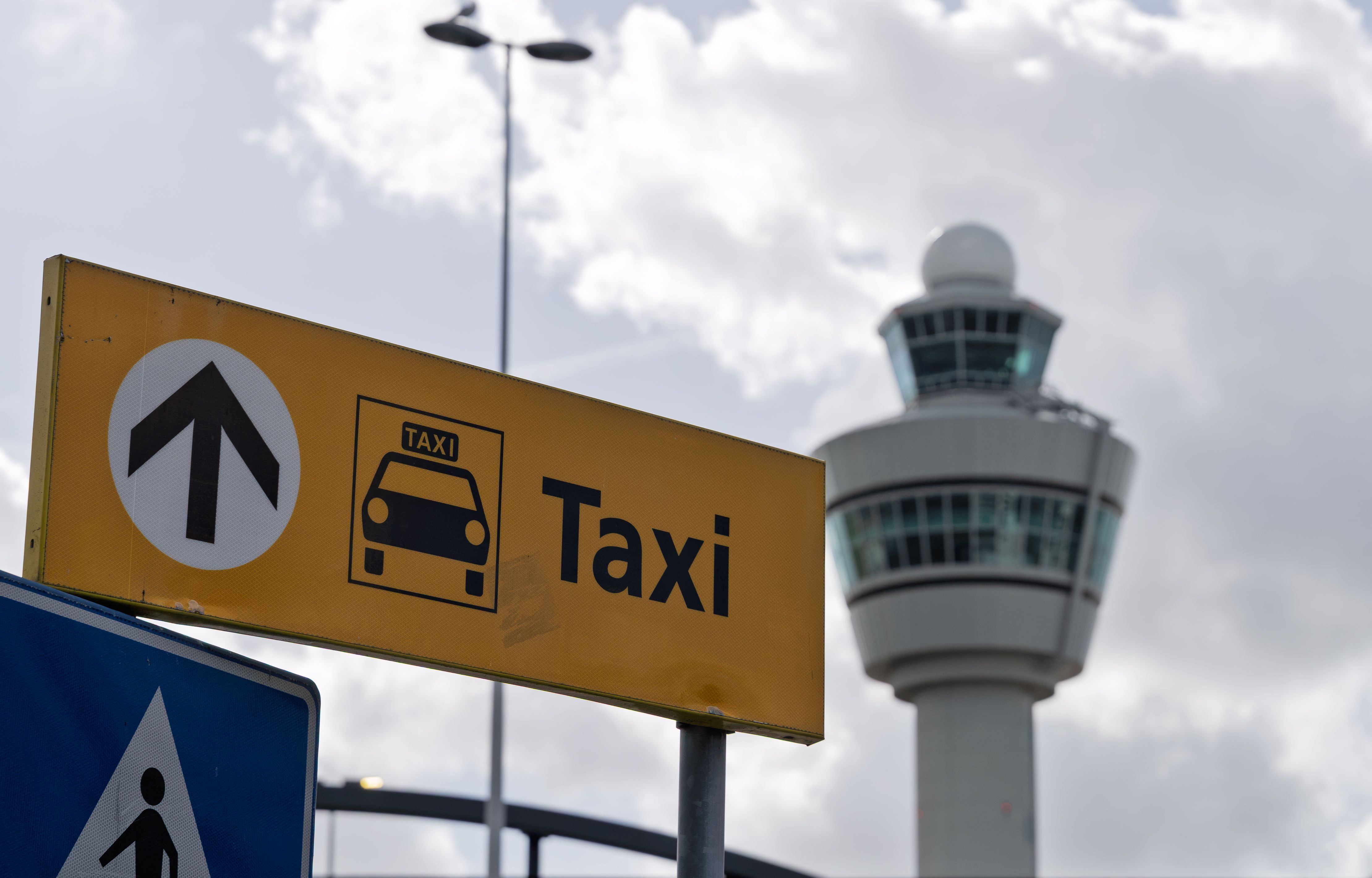 Yellow taxi sign with airport control tower in the background under a cloudy sky.