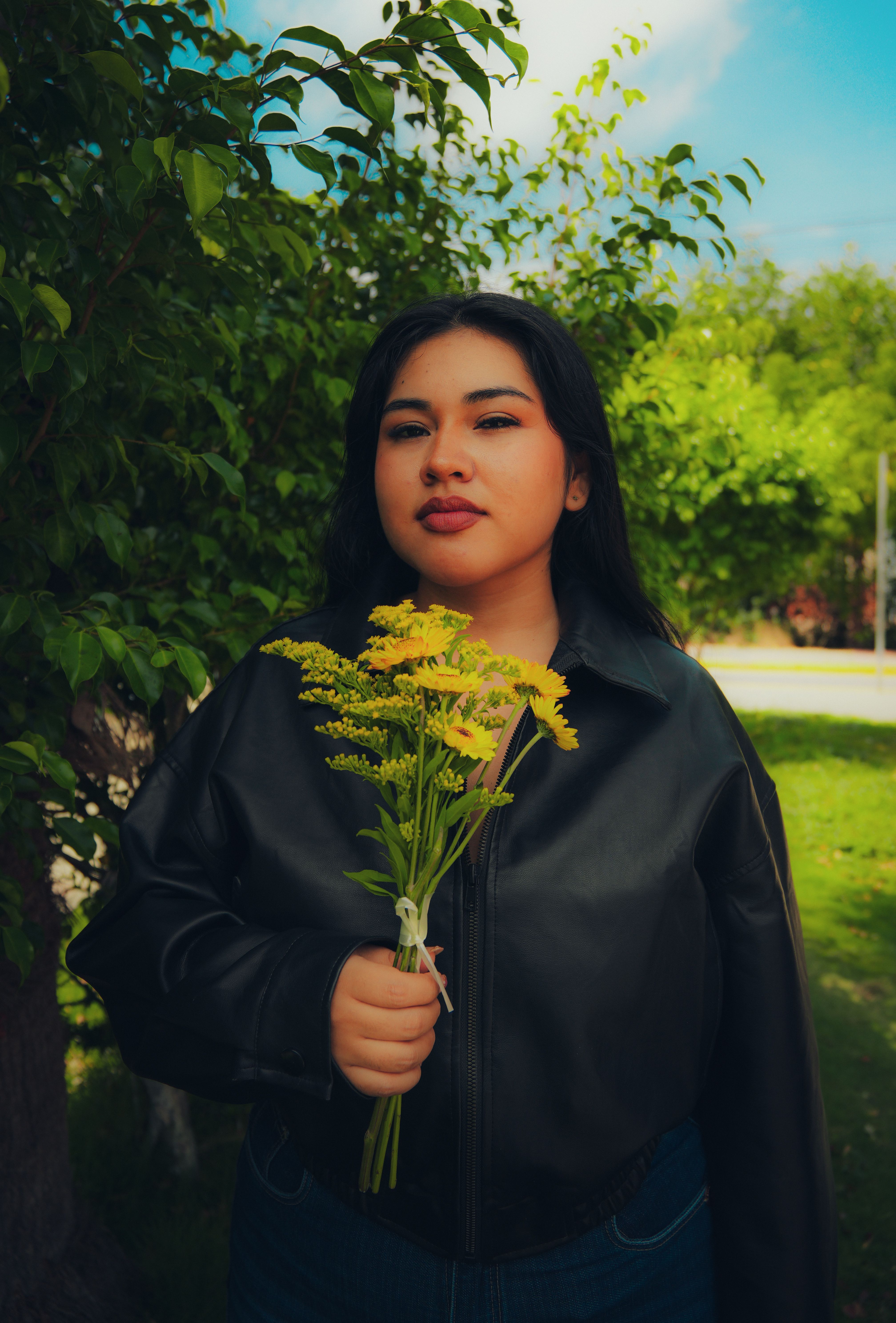 Young woman in a black leather jacket holding yellow flowers outside on a sunny day.