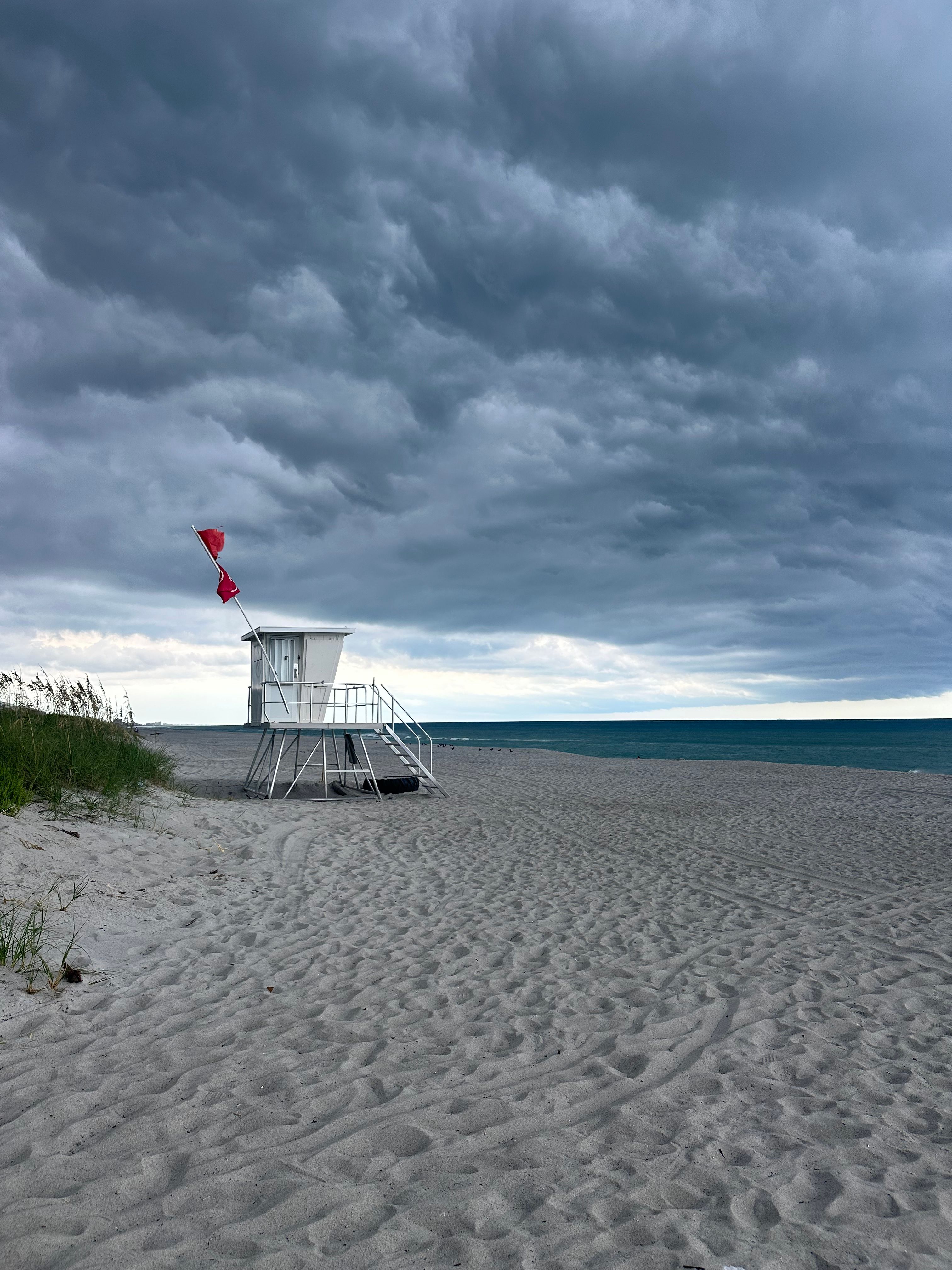 Dramatic storm clouds over a sandy beach with a lifeguard tower and red flag warning.