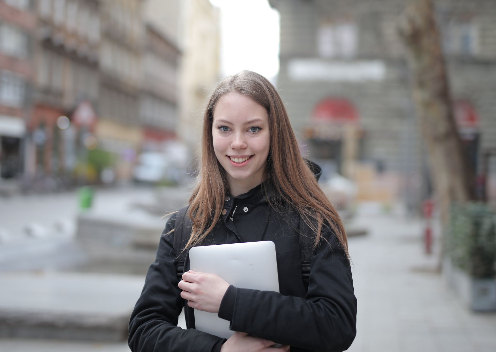 A young woman smiling while holding a laptop in an urban street setting, wearing casual attire.
