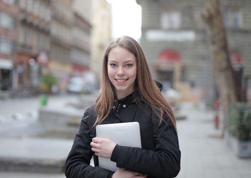 Free A young woman smiling while holding a laptop in an urban street setting, wearing casual attire. Stock Photo