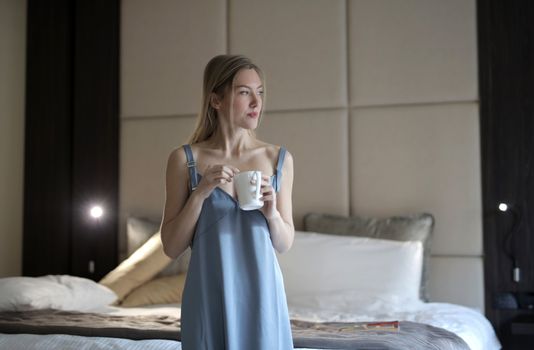 Woman enjoying morning coffee in blue sleepwear, standing in a cozy bedroom setting.