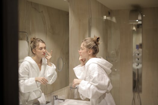 A woman in a white bathrobe applies moisturizer while looking at her reflection in the bathroom mirror.