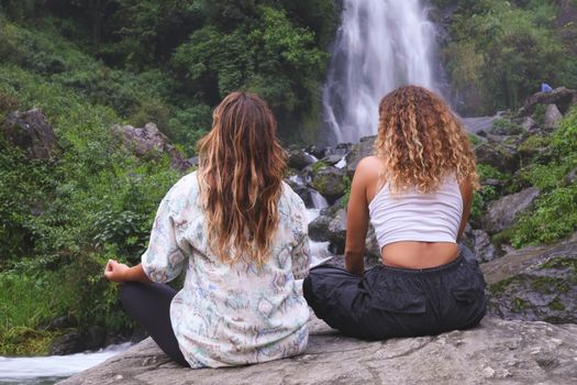 Two women practicing meditation near a stunning waterfall in a serene forest setting, captured from behind.