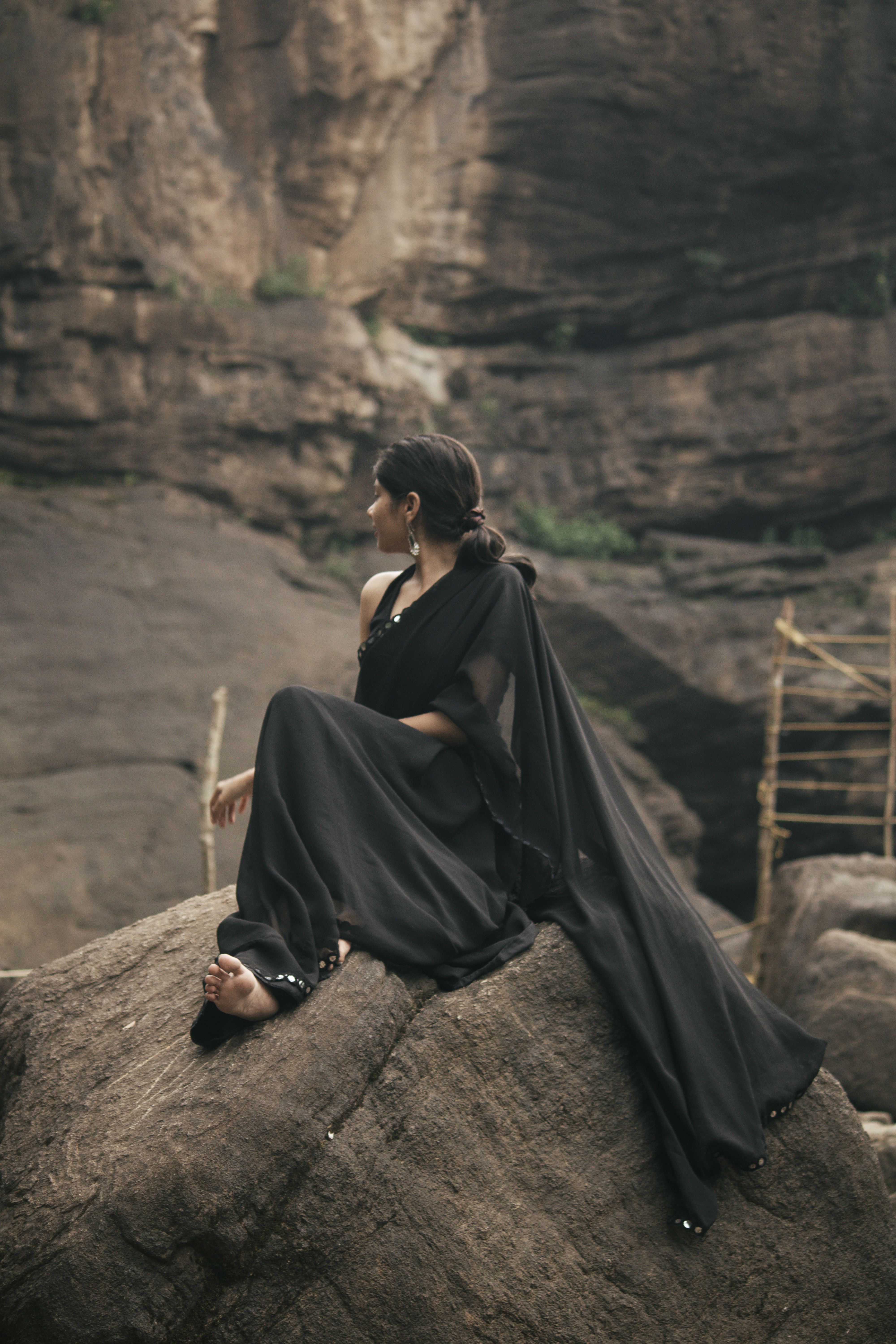 Fotomural Una mujer vestida con un sari negro se sienta con gracia sobre una roca en un entorno natural agreste.
