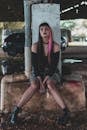 Young female goth with dark makeup in stylish clothes with bare shoulders sitting on concrete pedestal and looking at camera in covered parking on daytime
