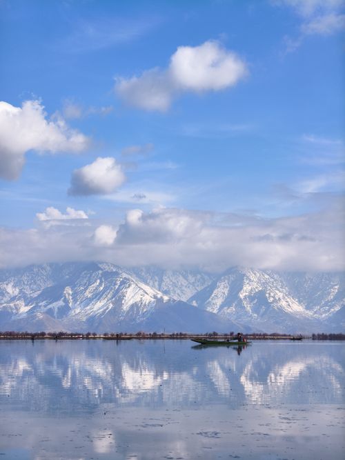 Free Traditional boat on a tranquil lake reflecting snow-capped mountains under a bright blue sky. Stock Photo