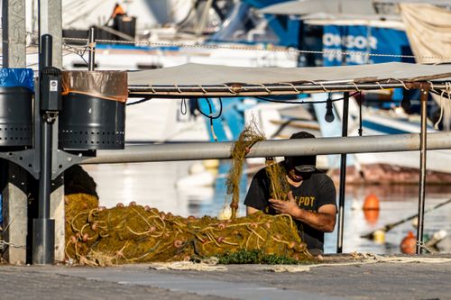 Free A fisherman repairs his net at a bustling harbor, surrounded by boats and marine equipment. Stock Photo
