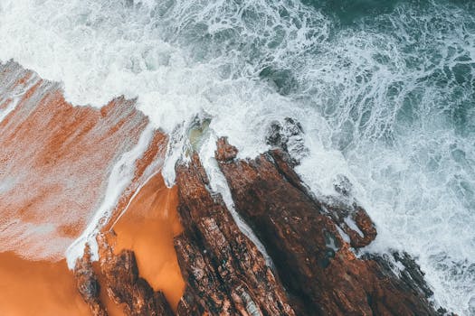 Drone shot capturing waves crashing onto a rocky coastline with vibrant orange sand.