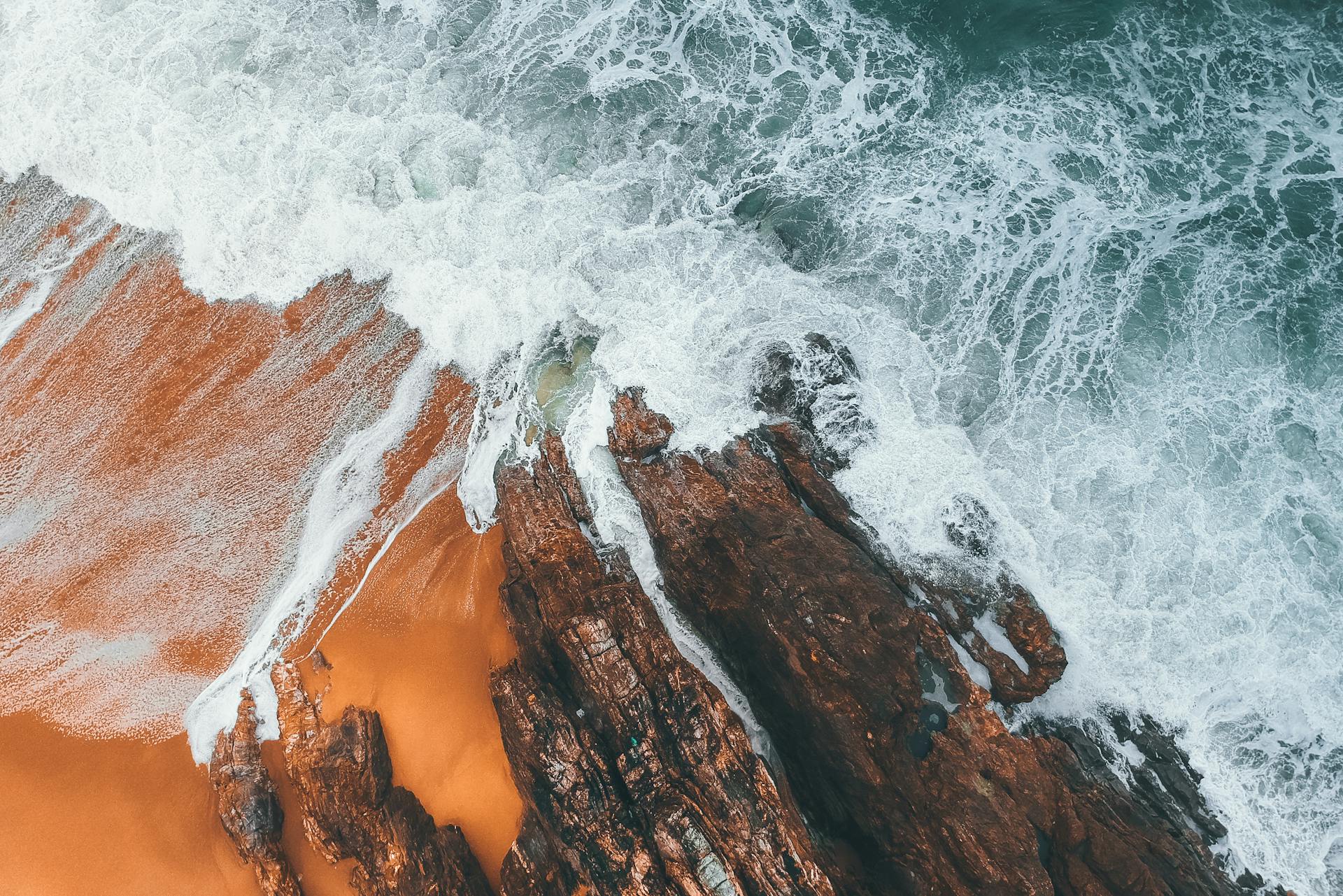 Drone shot capturing waves crashing onto a rocky coastline with vibrant orange sand.