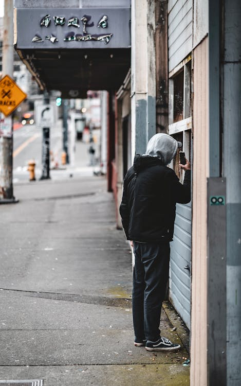 Back view of anonymous guy with smartphone in casual clothes with hood standing near building and looking at window while strolling on sidewalk of modern city street