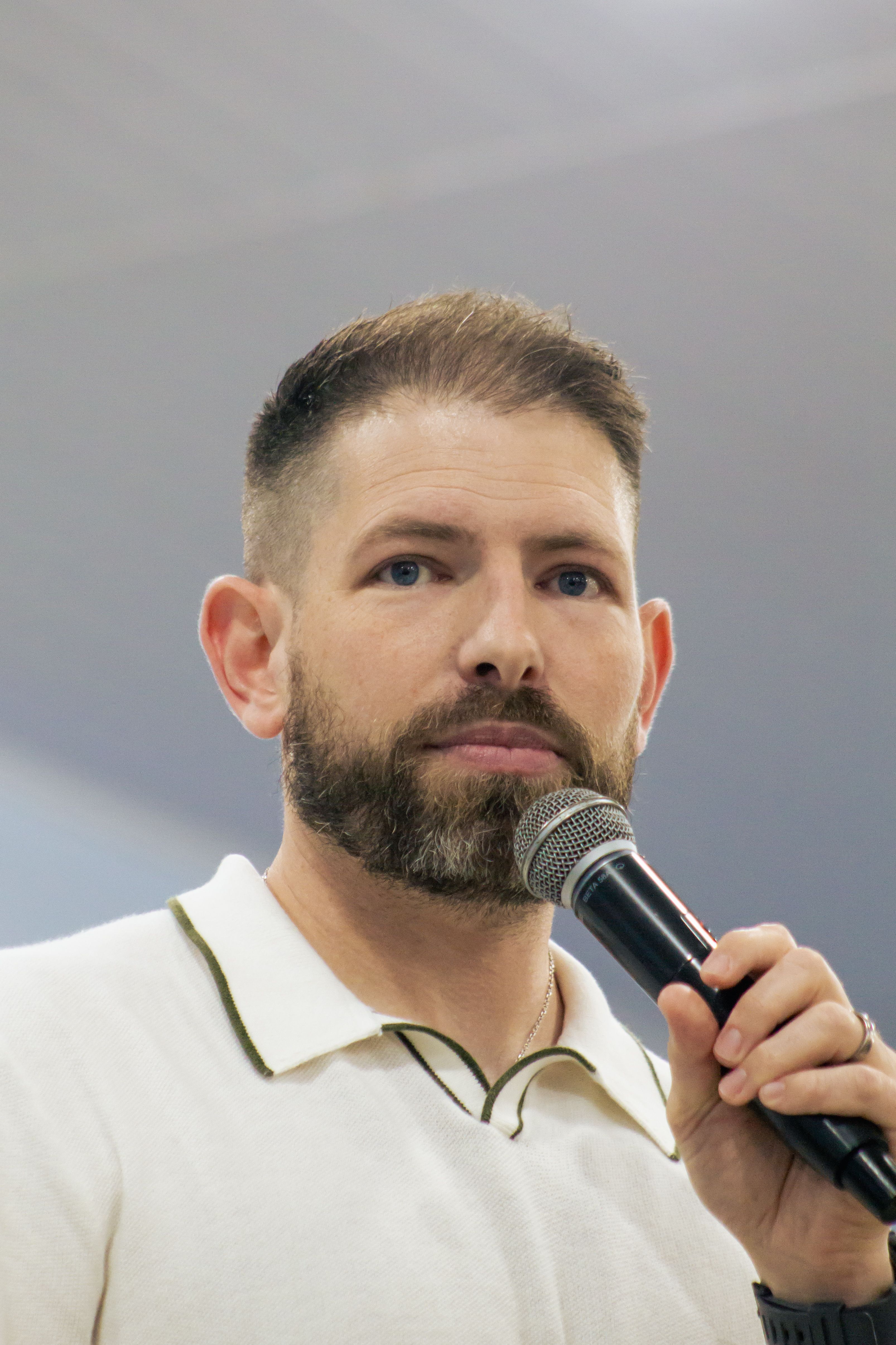Portrait of a bearded man speaking into a microphone, wearing a white shirt indoors.