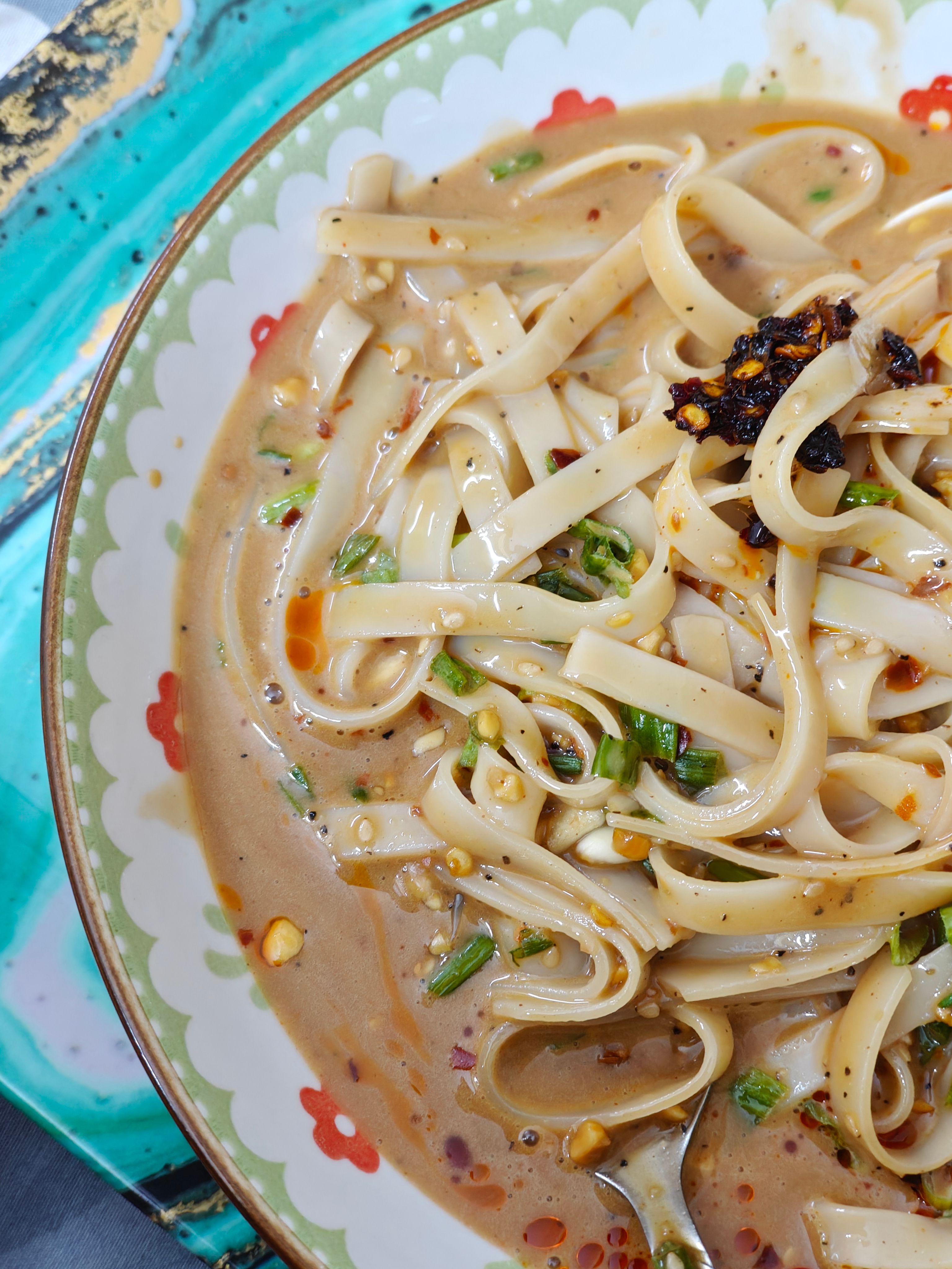 A small white bowl of glossy tan peanut sauce with a drizzle swirled on top, chopped peanuts and cilantro scattered around, and a spoon resting in the bowl.