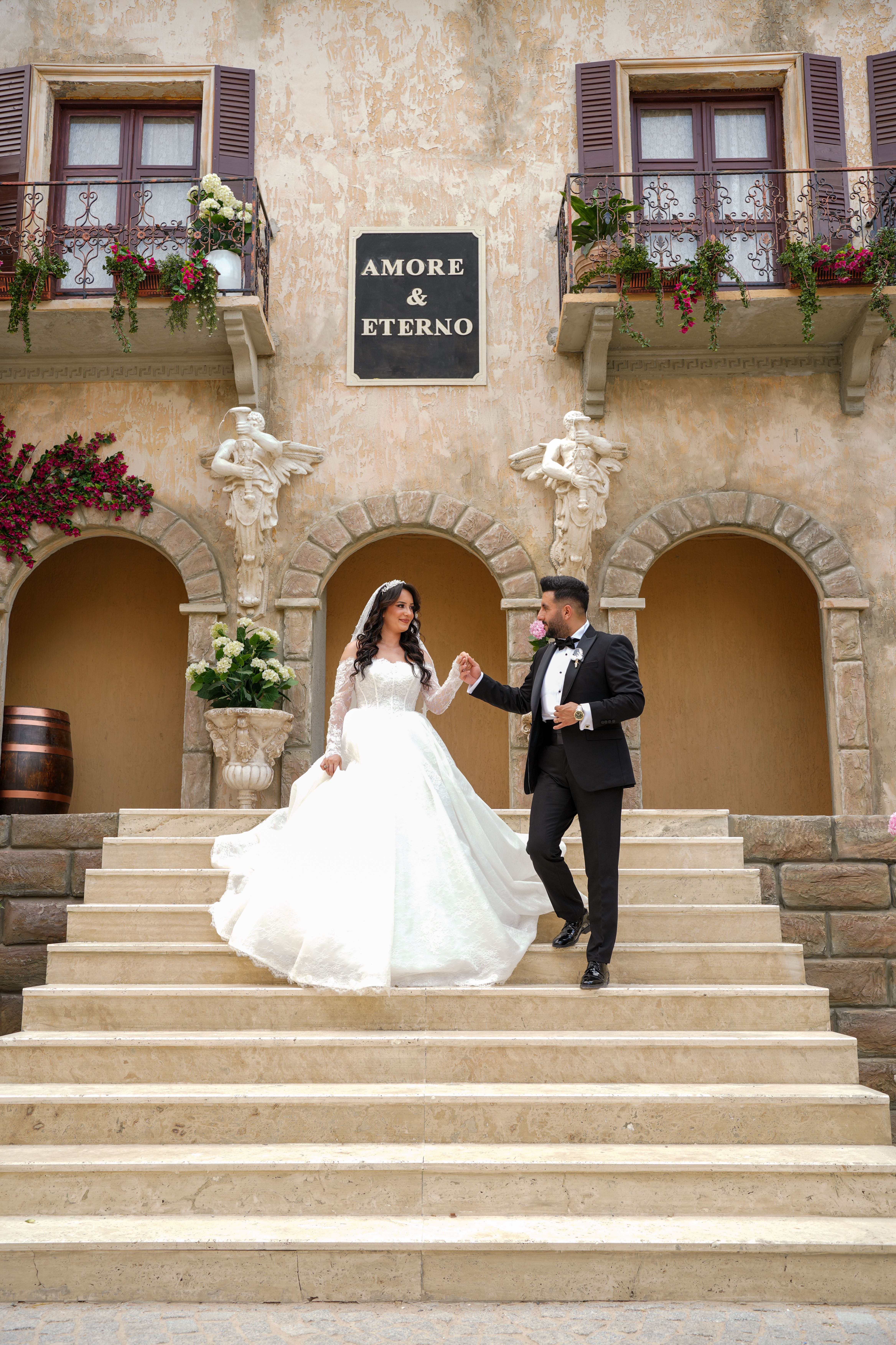 Bride and groom elegantly pose on a grand staircase of a historic building with 'Amore & Eterno' sign.