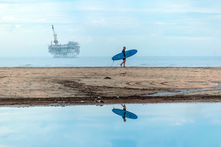 A lone surfer walks along a beach, carrying a surfboard, with an offshore oil rig in the background.