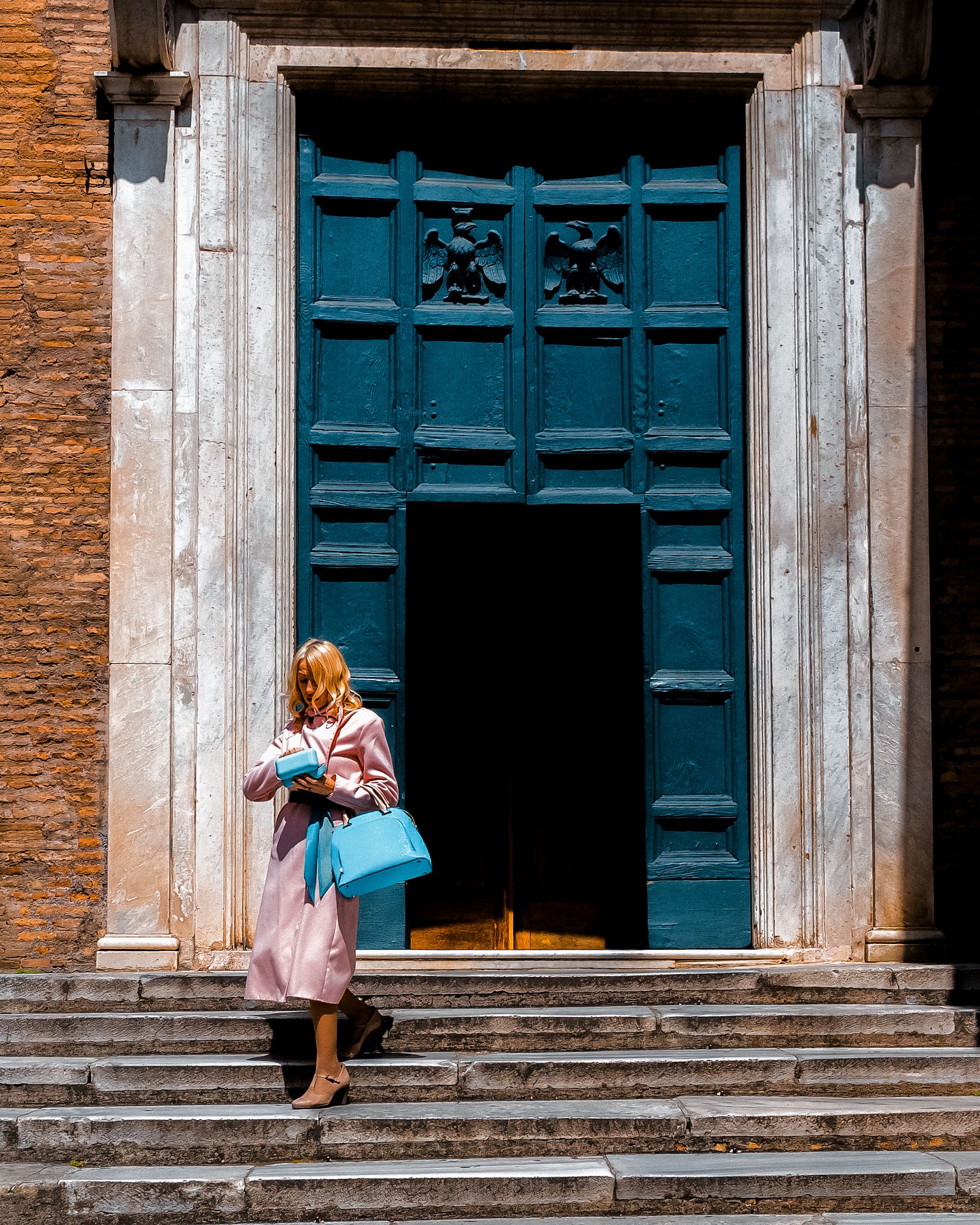 A stylish woman in Rome steps out of a grand blue doorway, showcasing urban fashion.