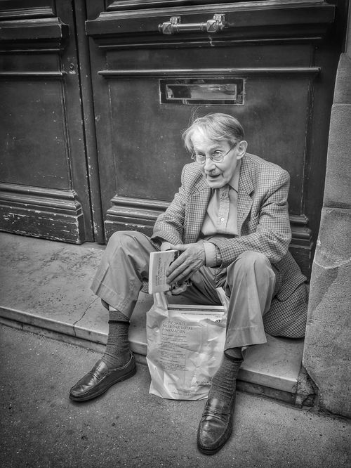 Free Black and white photo of an elderly man reading on a doorstep, showing solitude and contemplation. Stock Photo