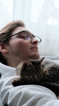 A young man in glasses relaxes indoors with a tabby cat resting on his chest.