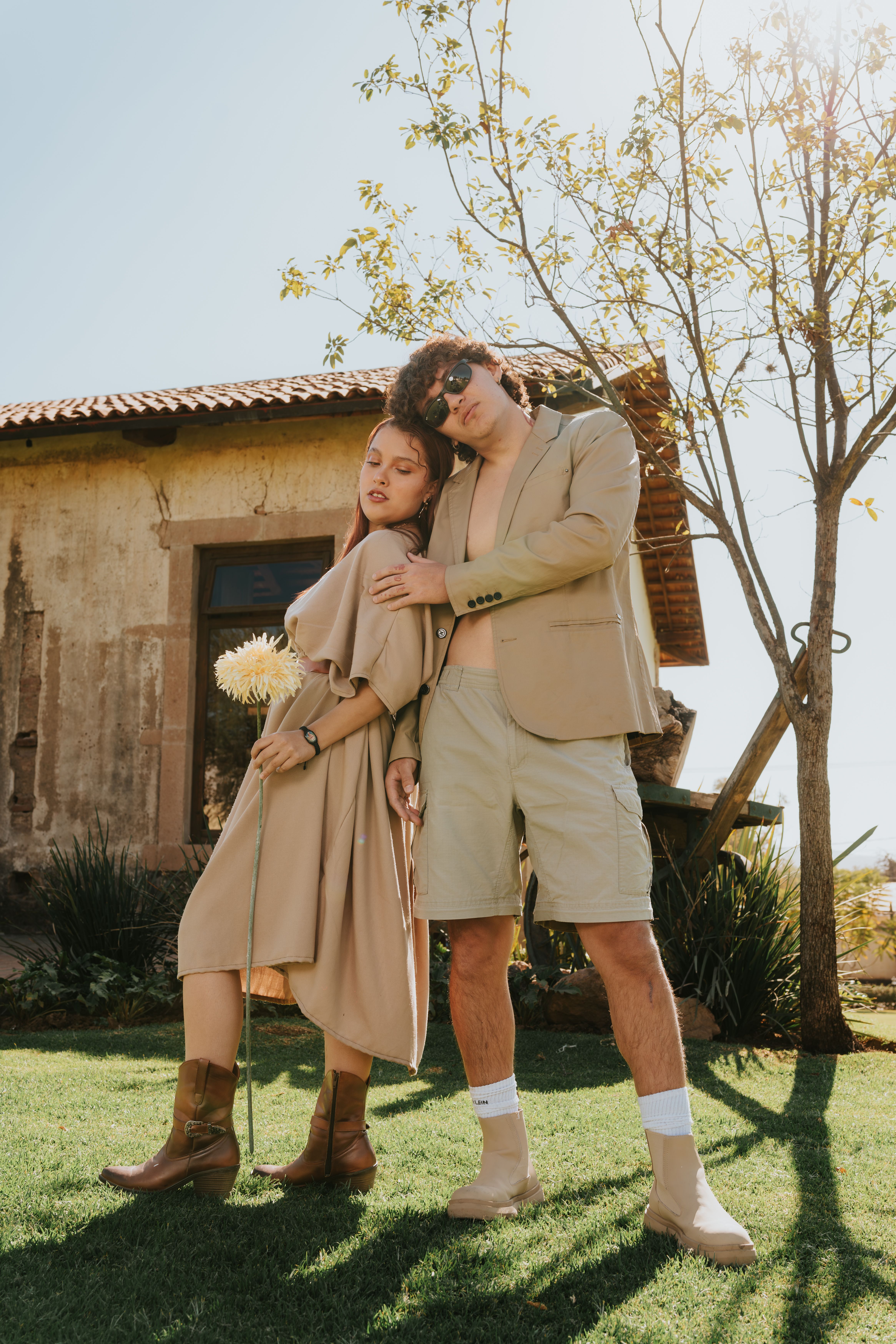 Stylish couple in beige outfits posing by rustic house under clear blue sky.
