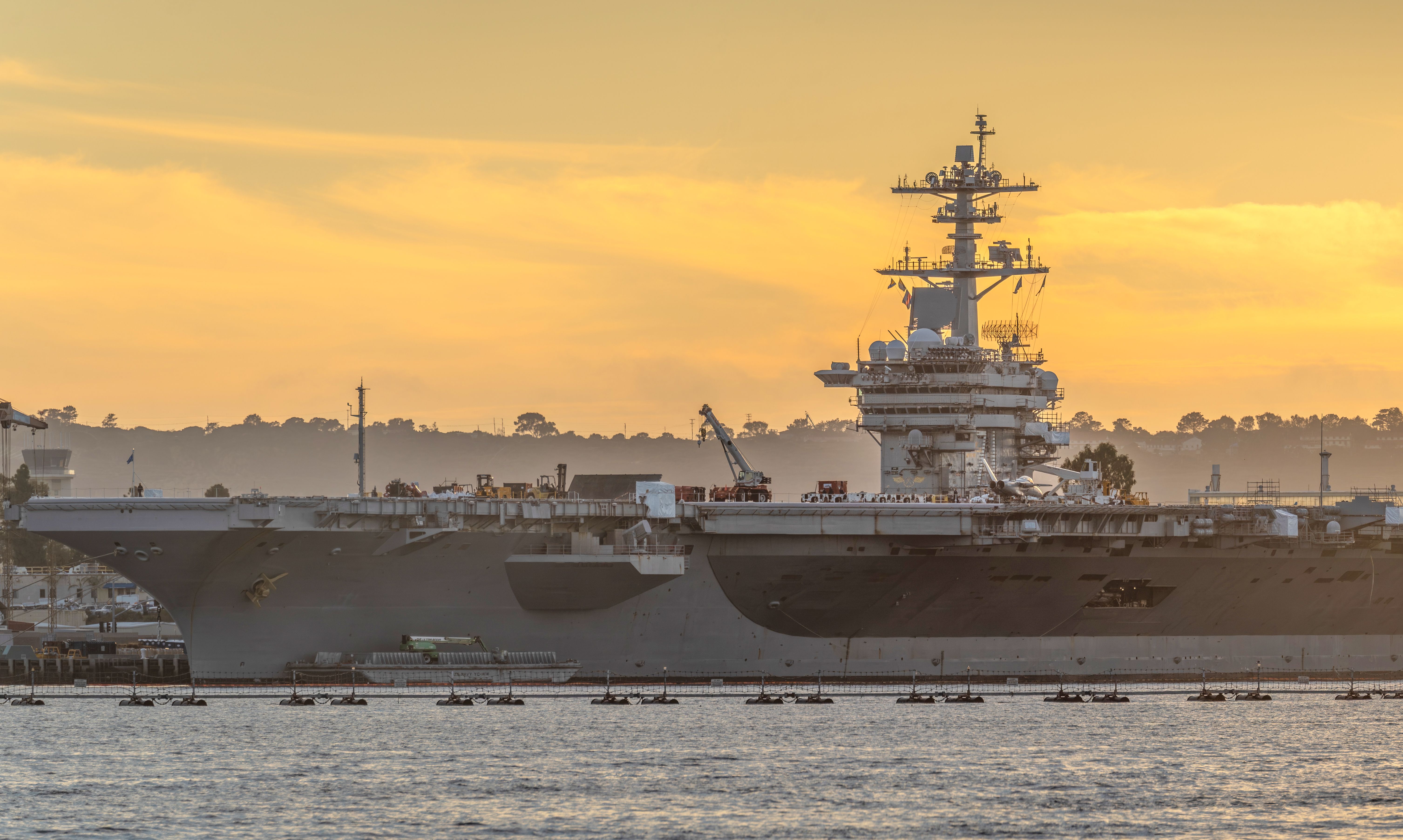 Large aircraft carrier docked at a harbor during a vibrant sunset, creating a dramatic nautical scene.