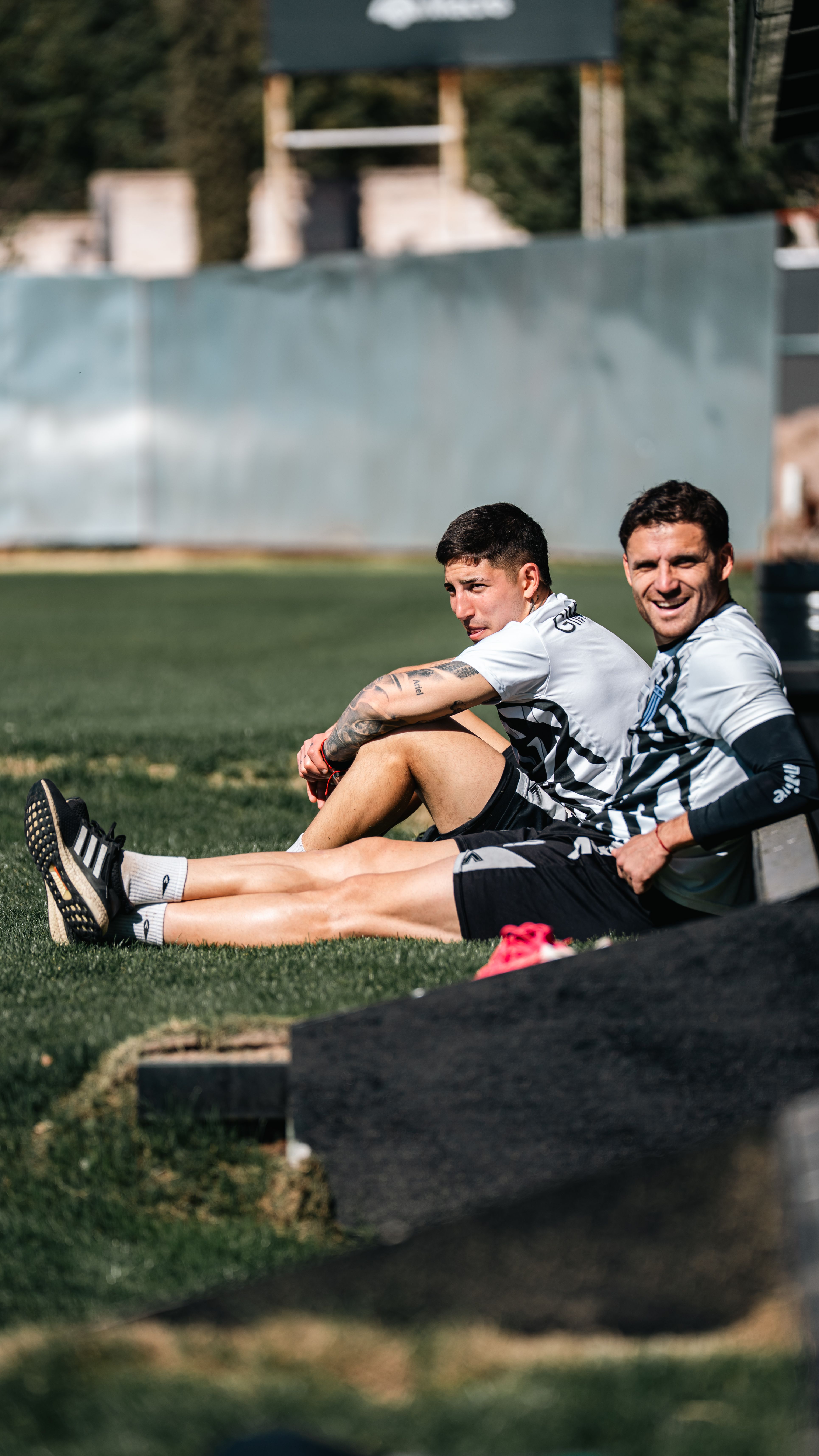 Two men sitting on grass after a game, enjoying a sunny day outdoors on a football field.