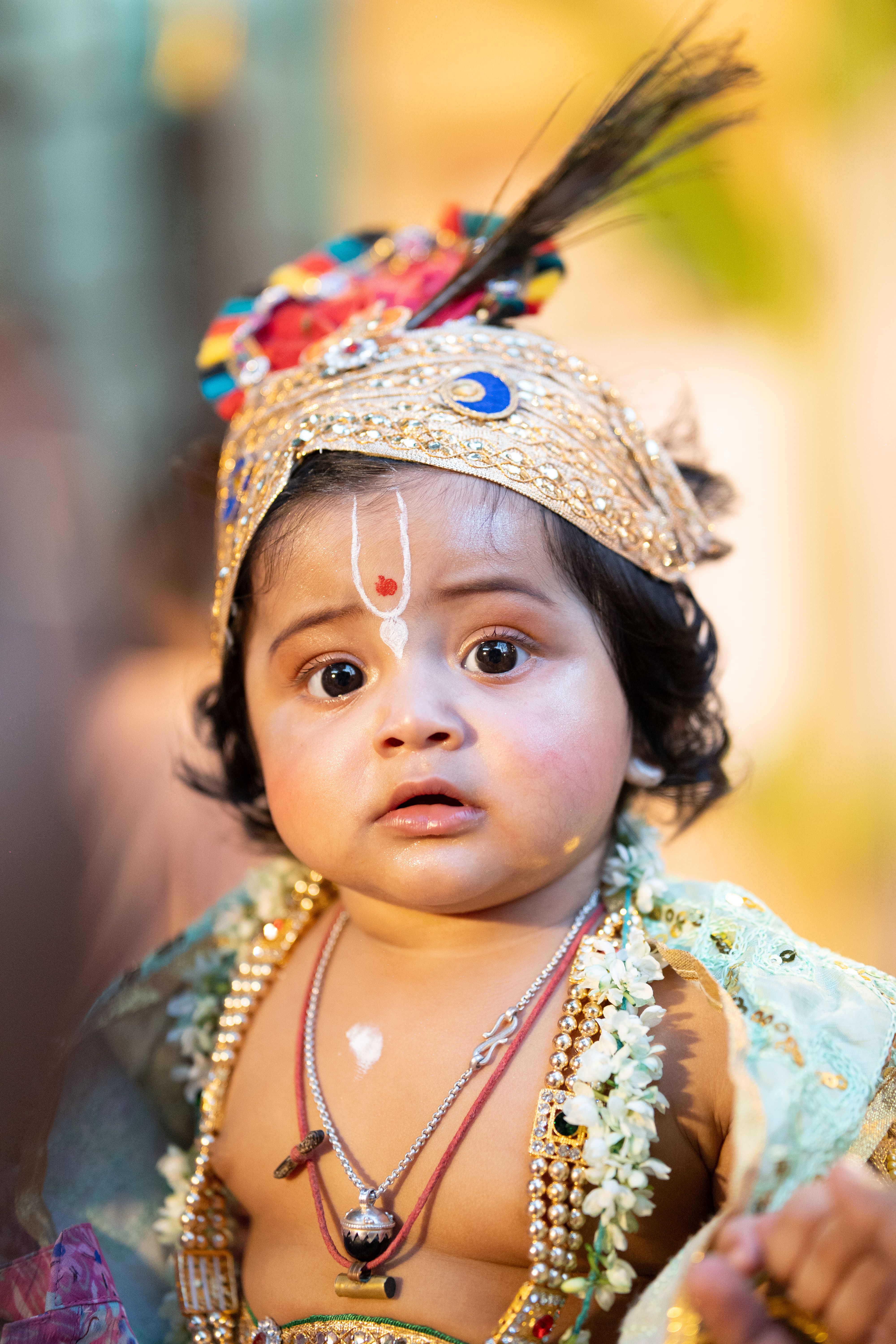 Adorable baby dressed in traditional attire with ornate headpiece, looking surprised.