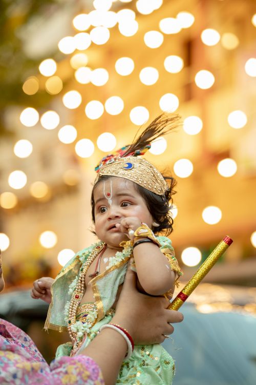 Free A cute baby dressed as Krishna, adorned with traditional attire, aglow with festive bokeh lights in the background. Stock Photo