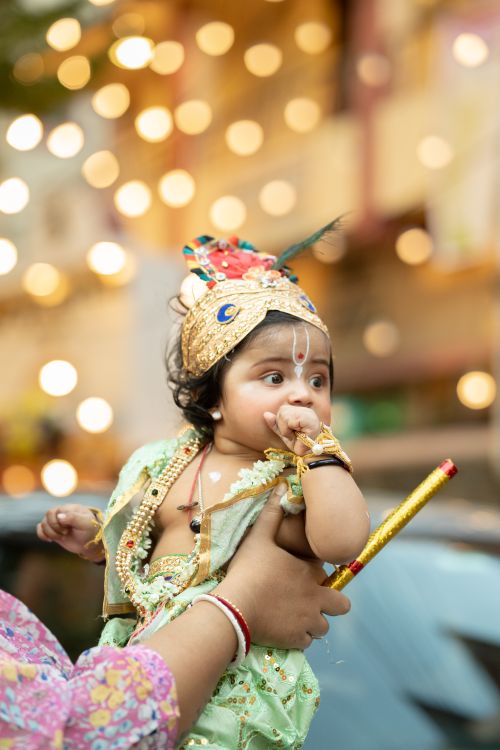 Free Baby dressed as Lord Krishna, celebrating Janmashtami with festive lights and traditional costume. Stock Photo