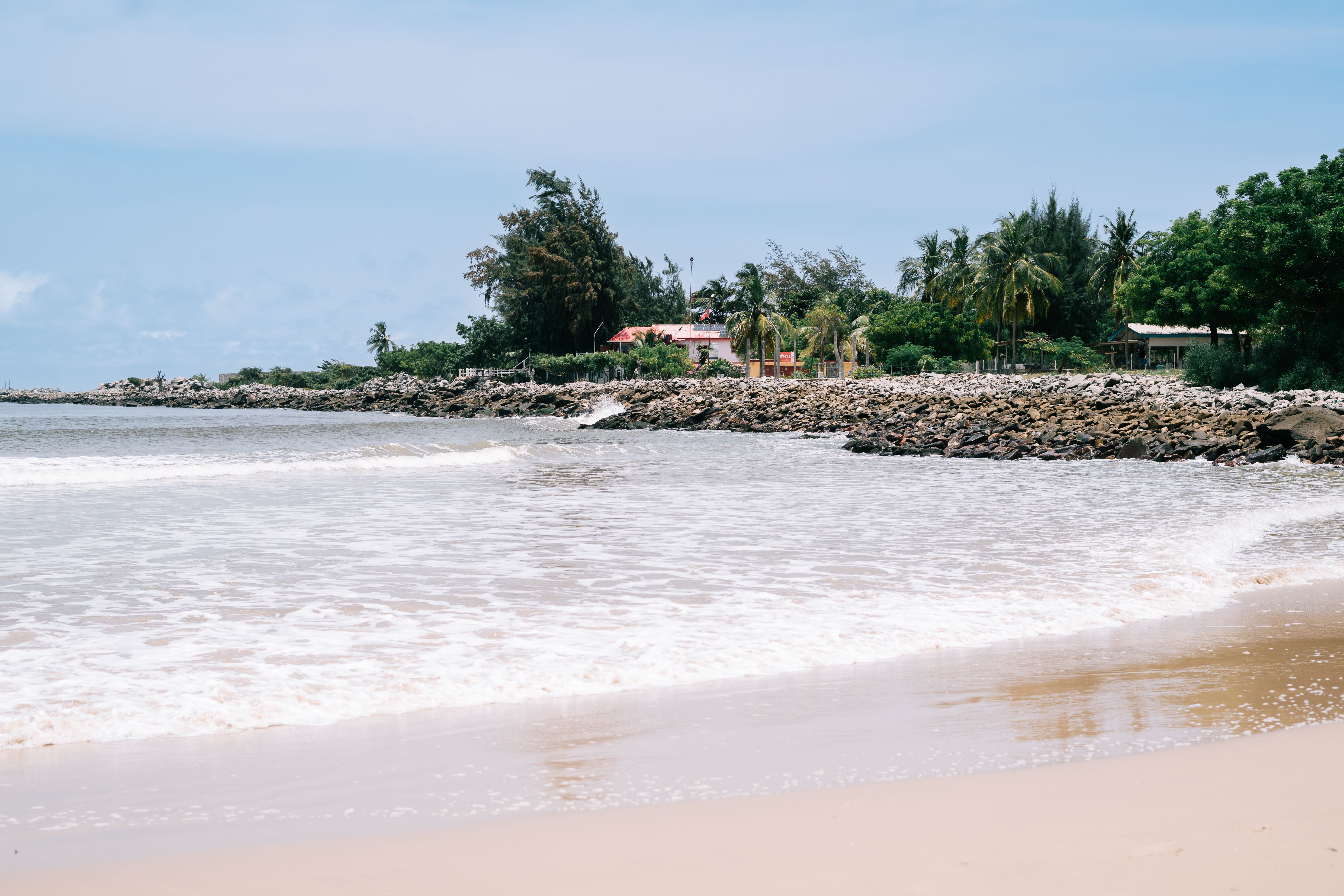 A serene tropical beach scene with gentle waves, palm trees, and a rocky shoreline under a clear blue sky.