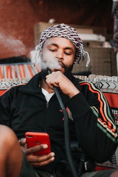 Serious ethnic guy in casual clothes and traditional headdress browsing smartphone and smoking hookah in modern apartment