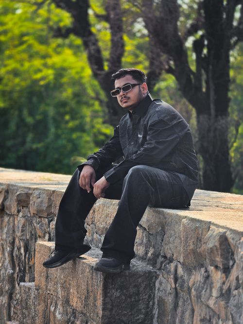Free Young man in dark attire sitting on stone wall outdoors with sunglasses, exuding a cool vibe. Stock Photo