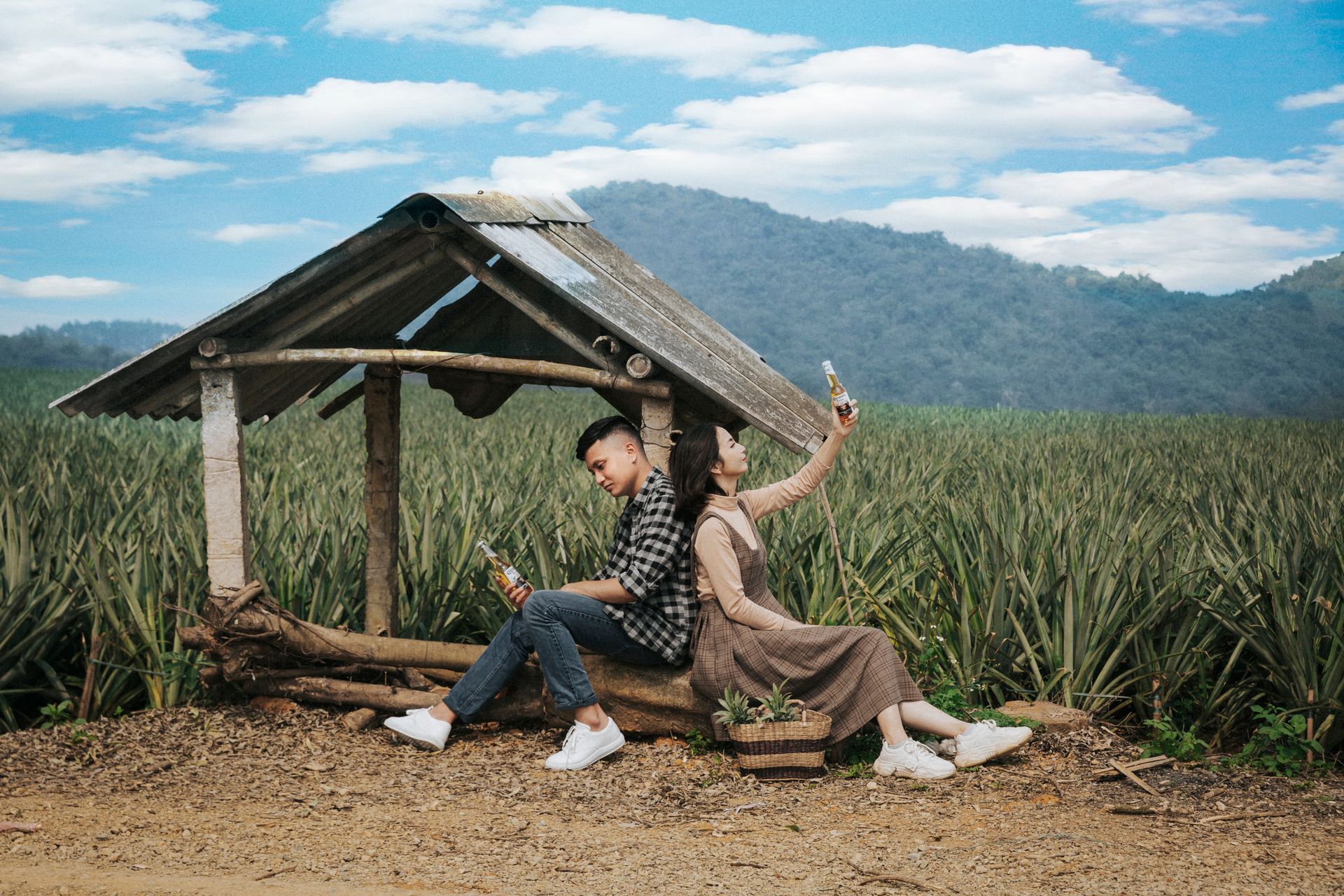Side view of young Asian man and woman in casual clothes sitting together back to back in dilapidated gazebo with glass bottles of drink in green field on sunny day