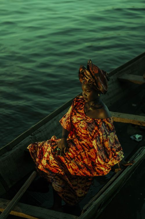 Free A woman in traditional attire sits on a canoe at twilight, surrounded by water. Stock Photo