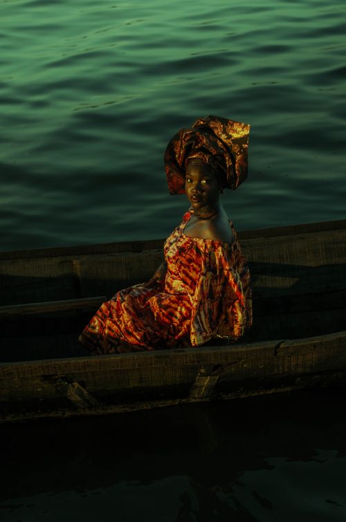 Free A woman in vibrant traditional attire sits on a canoe against a serene water backdrop at sunset. Stock Photo