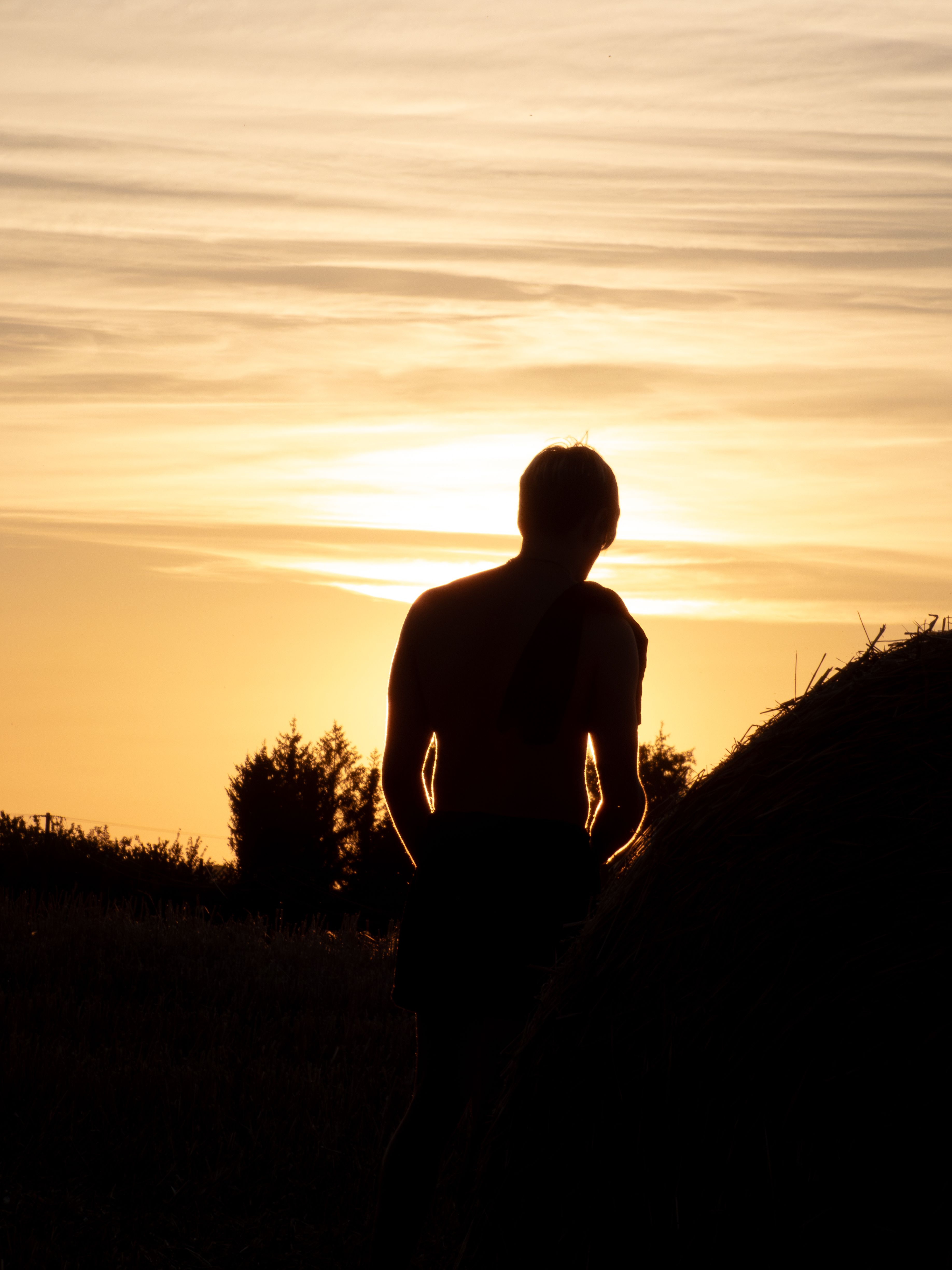 Fotomural La silueta de una persona de pie junto a una paca de heno durante una serena puesta de sol en un campo rural.
