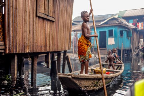 Free A child skillfully navigates a wooden canoe through a bustling lagoon village, highlighting daily life. Stock Photo