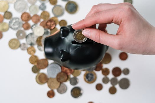 Close-up of a hand inserting a coin into a black piggy bank with scattered coins on a white background. ads can expose your pinterest posts , it can a good investment for your business