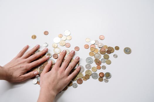 Hands arranging a variety of international coins on a neutral light background.