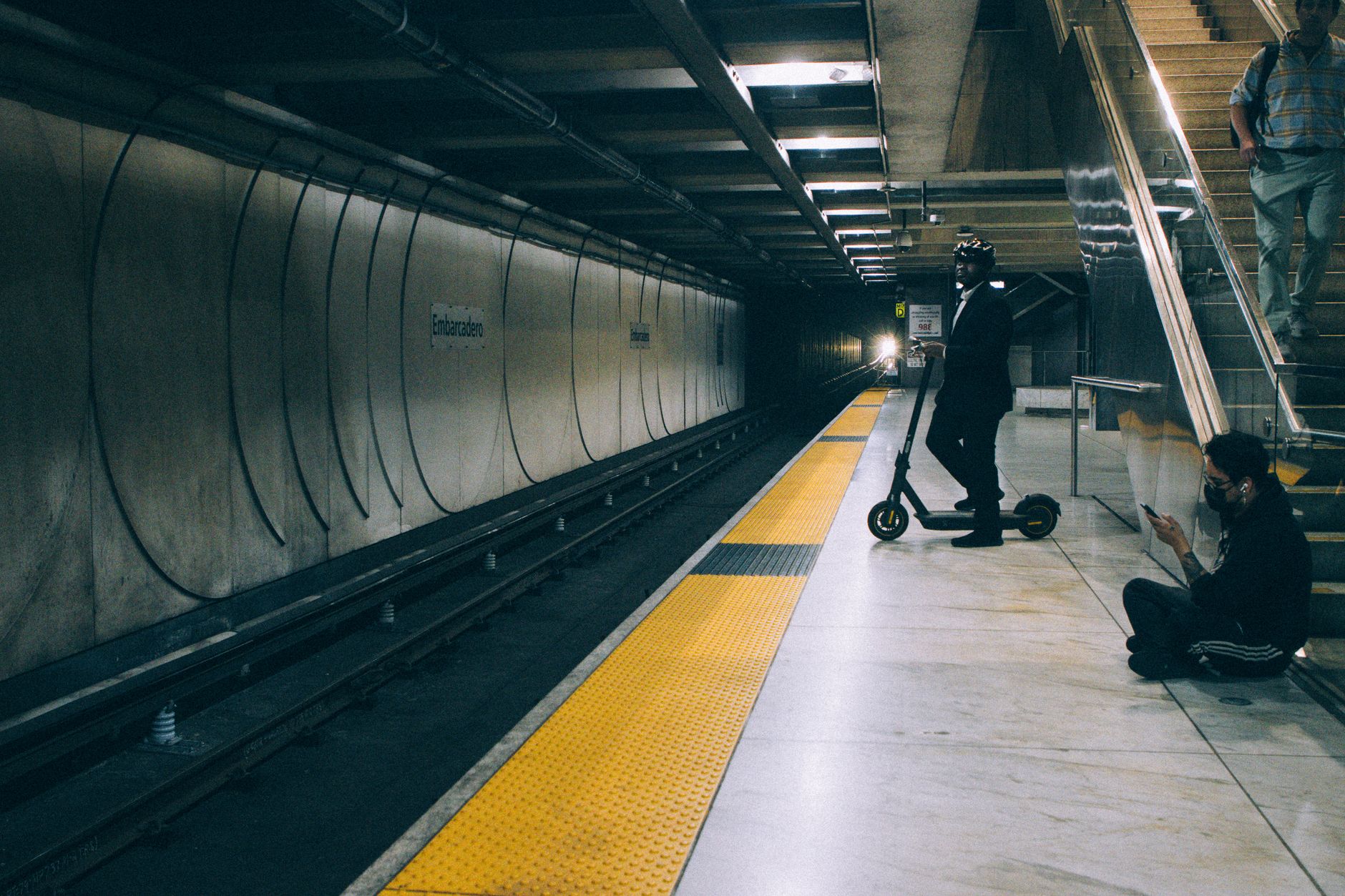 A commuter on a scooter waits at an Oakland subway platform as a train approaches in the distance.