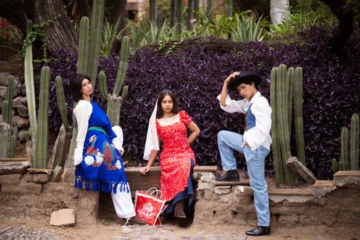 Three adults in traditional clothing posing in a cactus garden, showcasing vibrant Mexican fashion and culture.