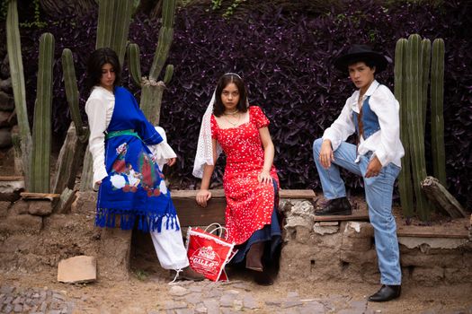 Three people in traditional Mexican attire posing outdoors with cacti.