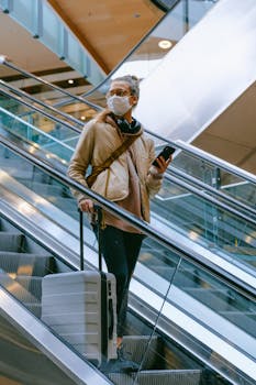 A woman wearing a mask uses an escalator in an airport with her luggage, reflecting modern travel norms.