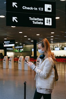 Woman wearing a mask, texting in airport terminal with signage in background.