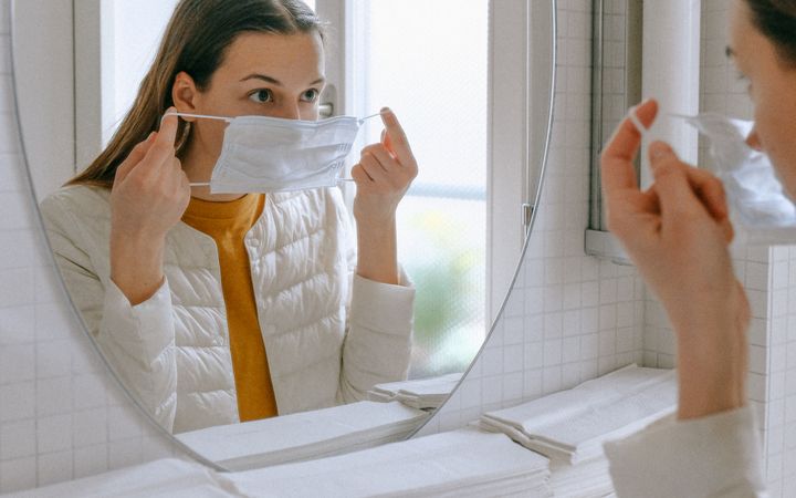 A woman adjusts her face mask in a bright bathroom mirror, practicing hygiene.