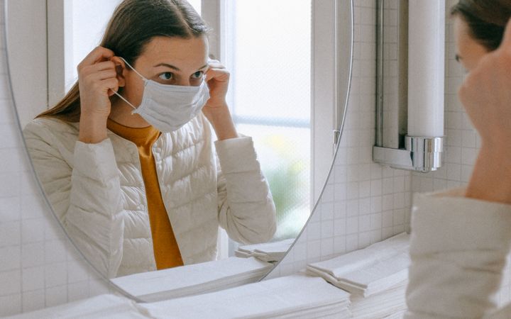 A woman adjusts her face mask while looking in the bathroom mirror, emphasizing hygiene and health.