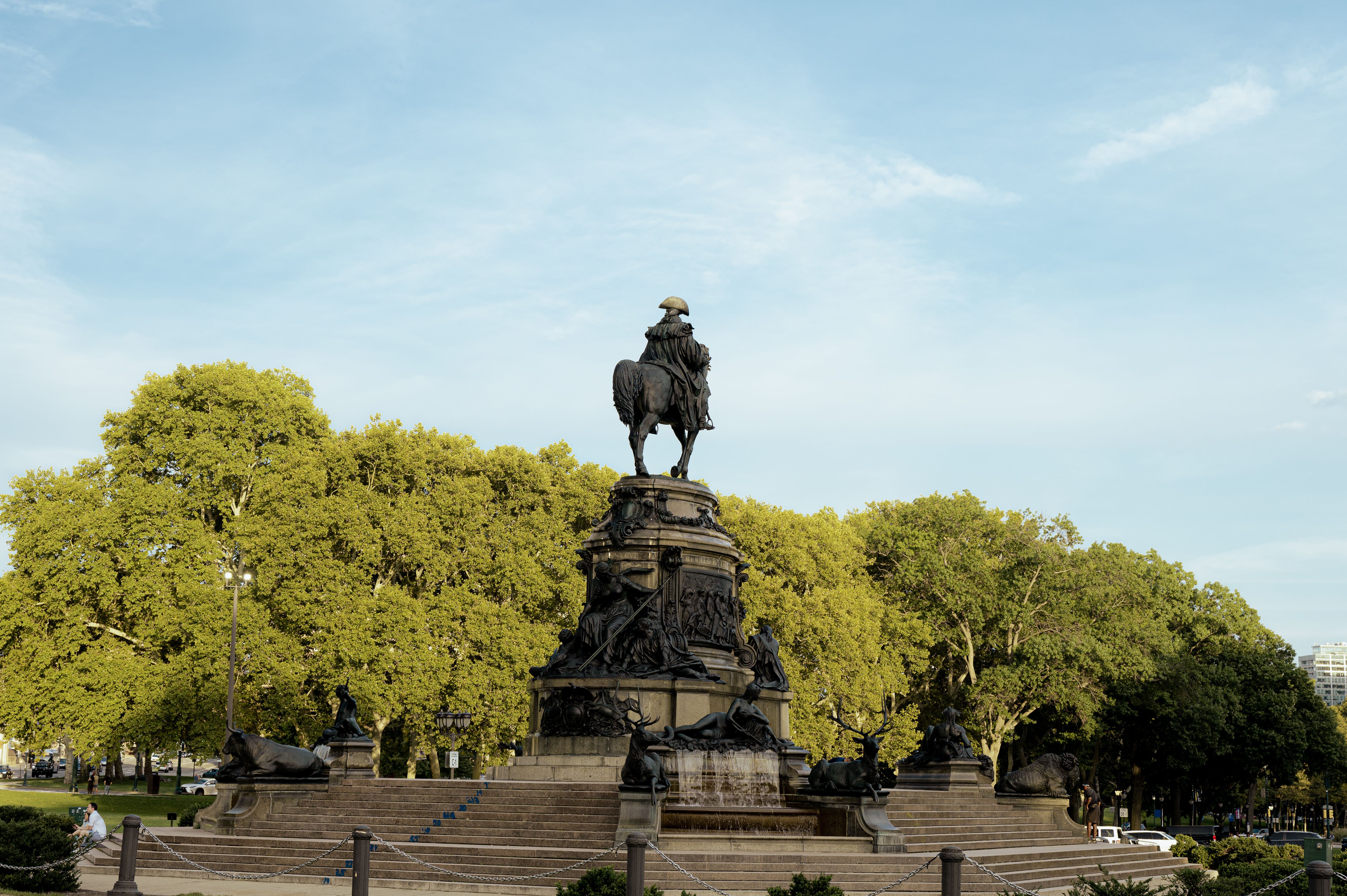 Bronze equestrian statue in city park surrounded by vibrant green trees under a clear sky.