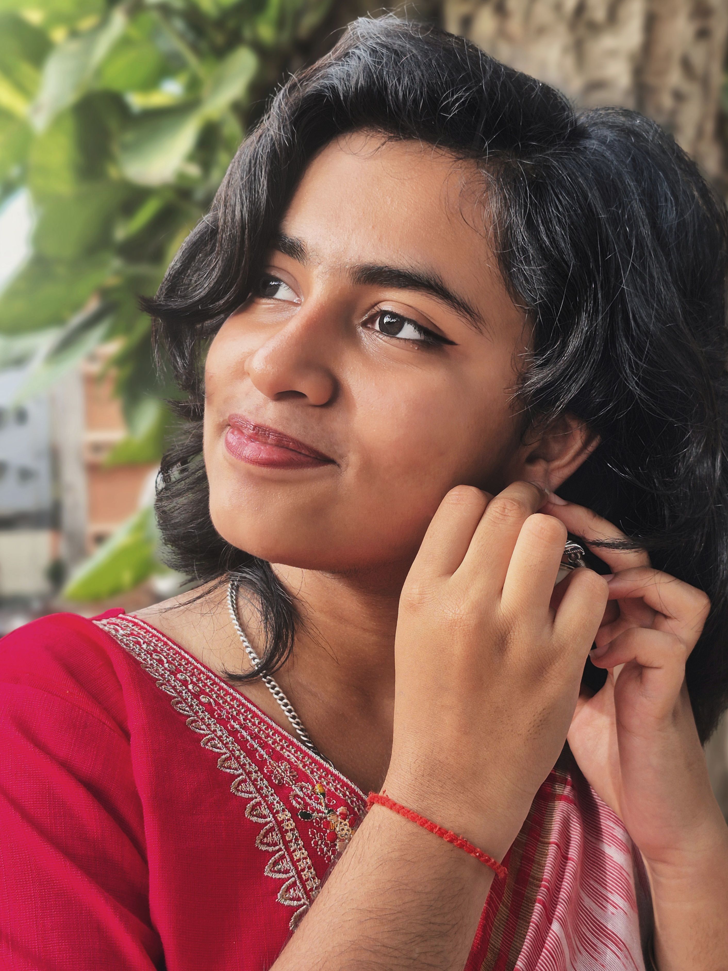 Portrait of a young woman adjusting her earring outdoors against a leafy backdrop, exuding elegance and confidence.