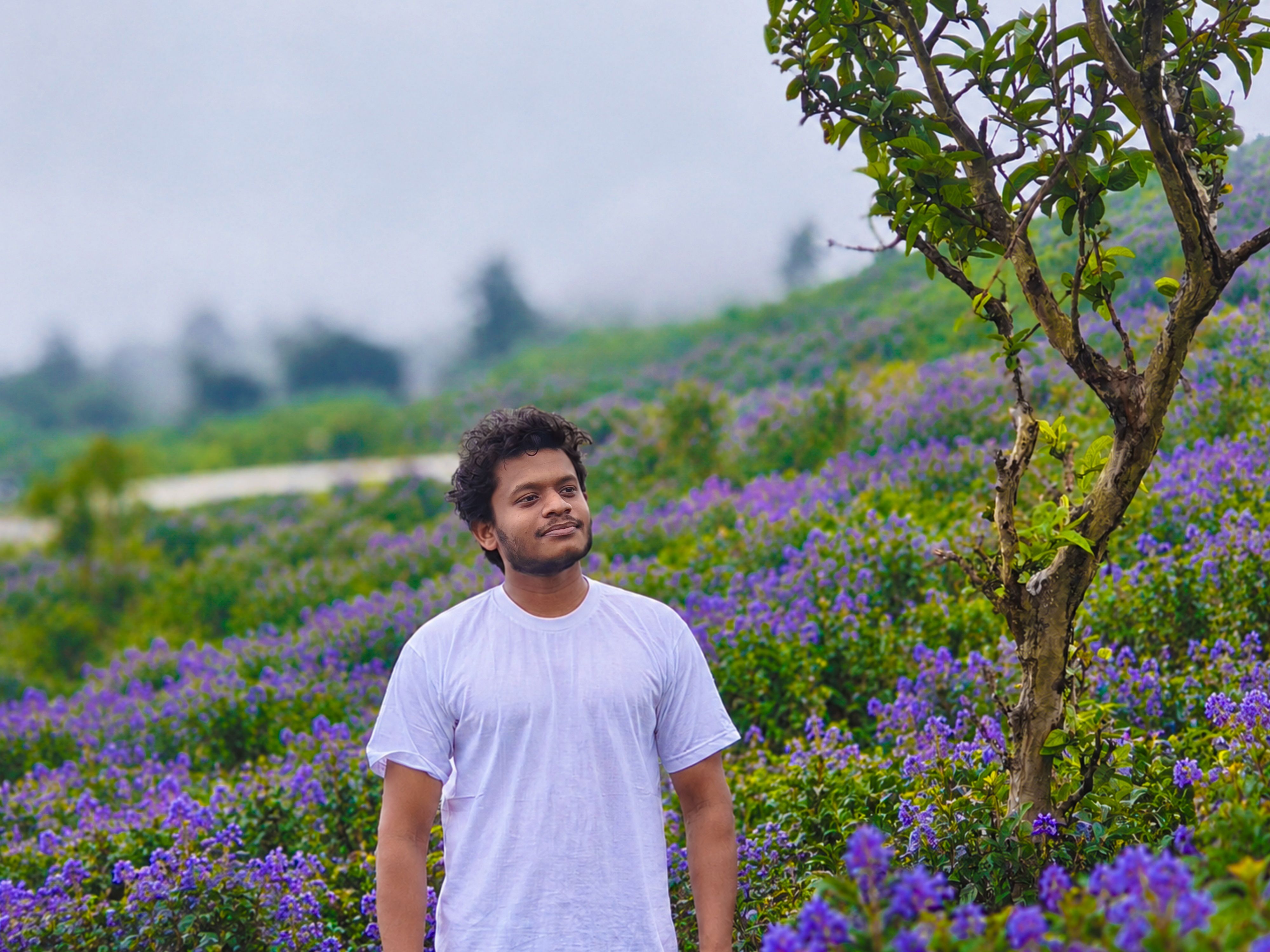 A serene portrait of a man in a white shirt standing in a lush field of purple flowers, under a cloudy sky.
