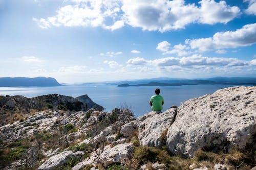 Free Man In Green Shirt Sitting On Rock Formation Near Sea Under Blue Sky Stock Photo